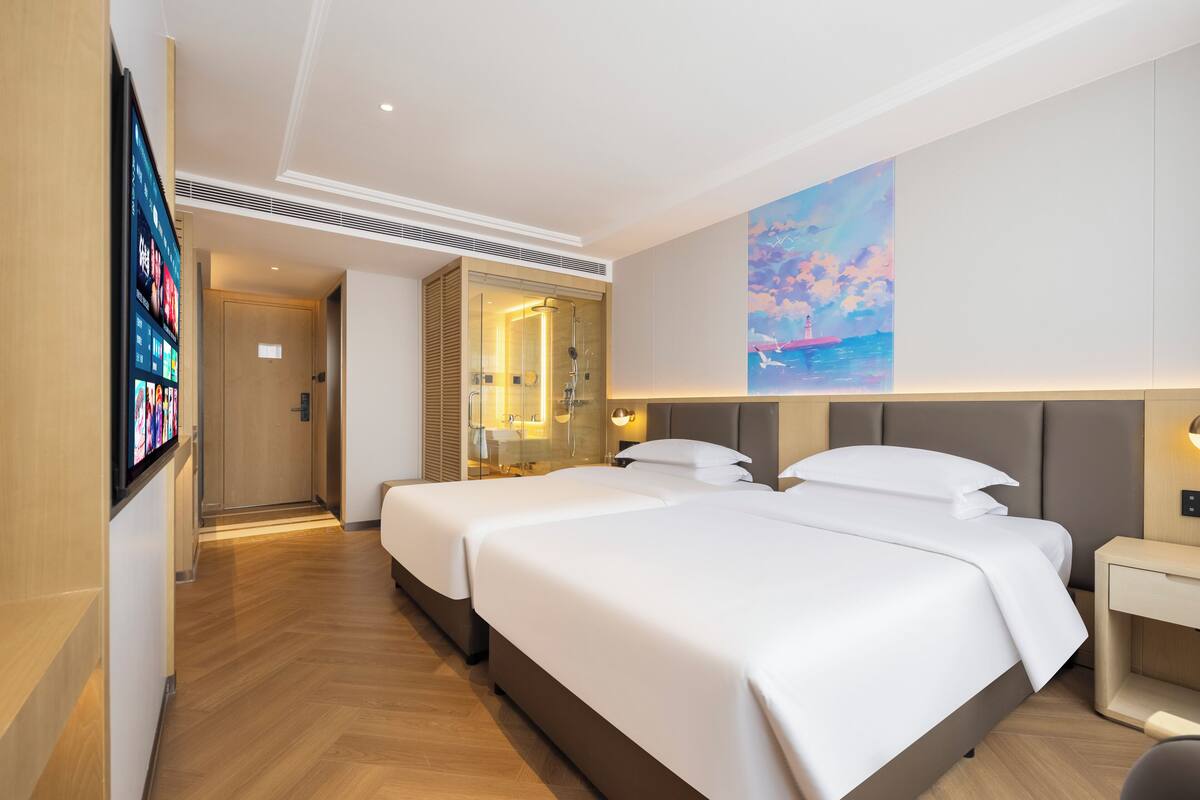 A hotel room with two beds, a TV, a painting, and a bathroom with a glass shower.