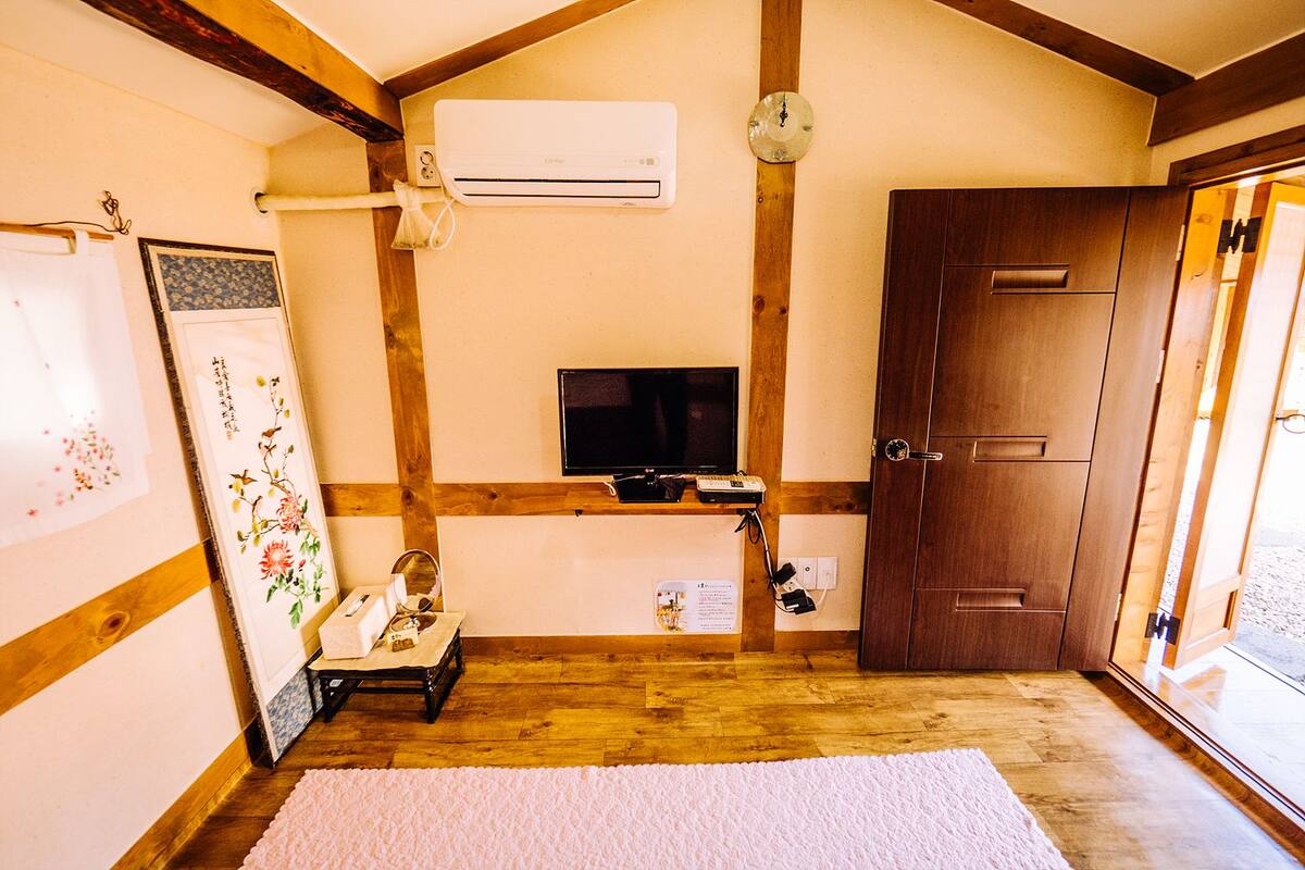 A room with wooden beams, a mounted air conditioner, a television on a shelf, a door, and a small table with a mirror and tissues.