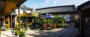 A traditional Japanese courtyard with a wooden table and chairs under a canopy, surrounded by potted plants and a gravel path. - Hohomom Guesthouse (Jeonju)