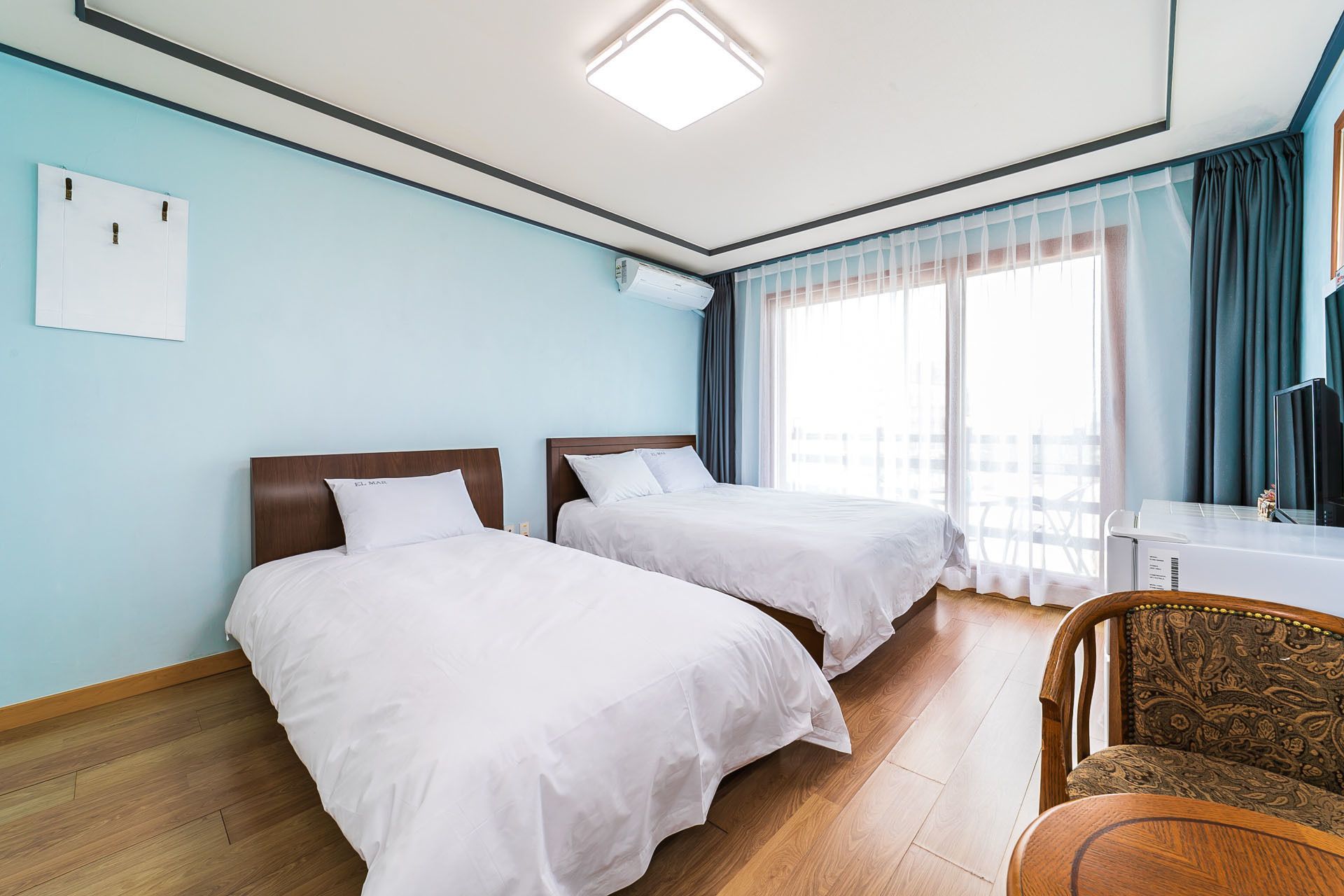 A hotel room with two beds, a desk, a chair, and a large window with curtains.