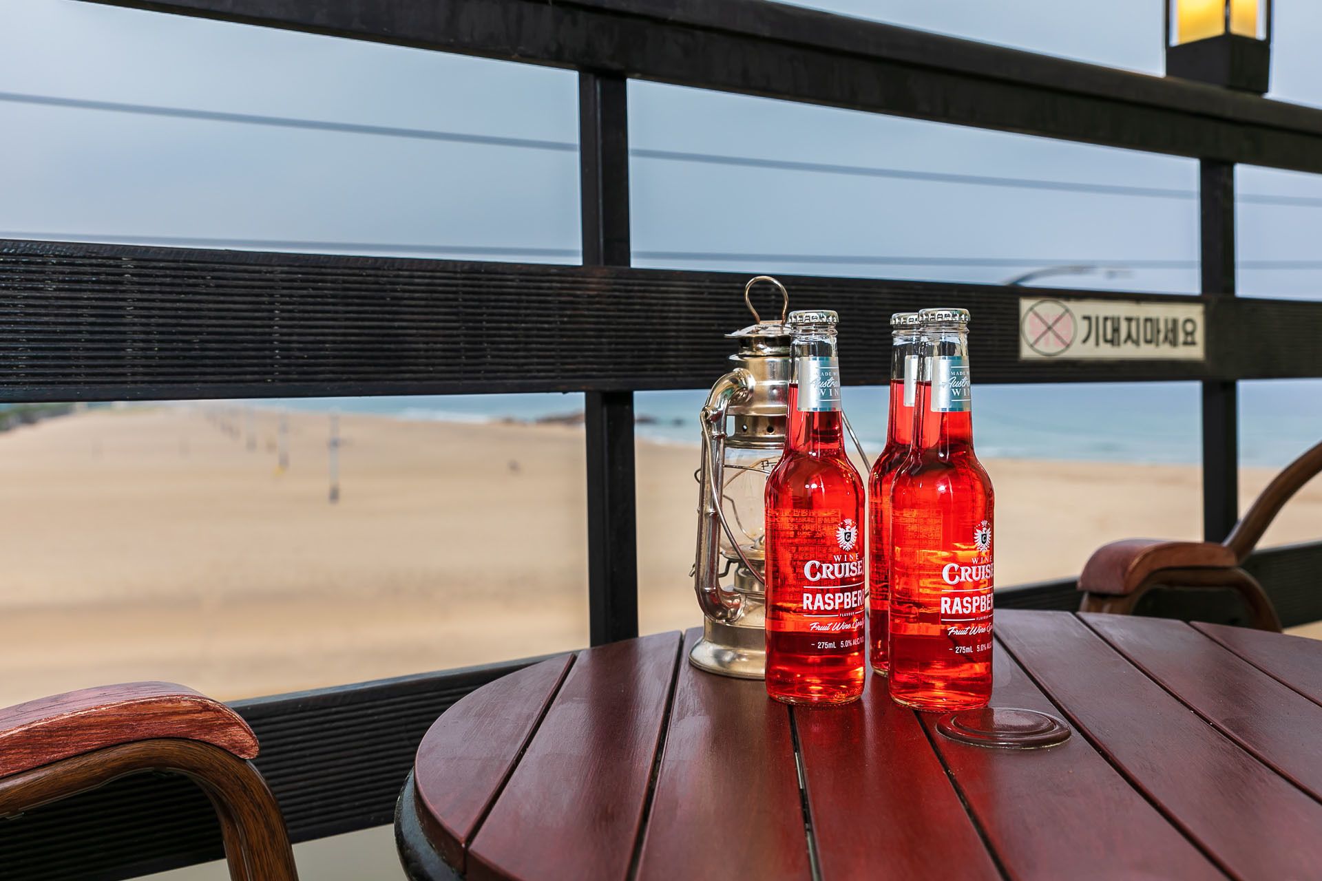 A table with two bottles of Raspberry wine and a metal lantern on a wooden deck overlooking a beach.