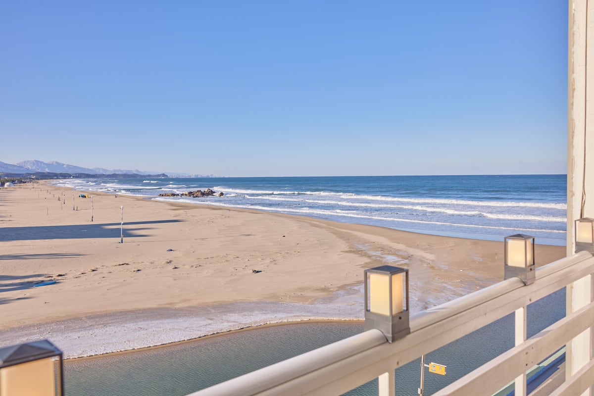 A beach view from a balcony with a railing and two lanterns, a sandy beach, and the ocean.