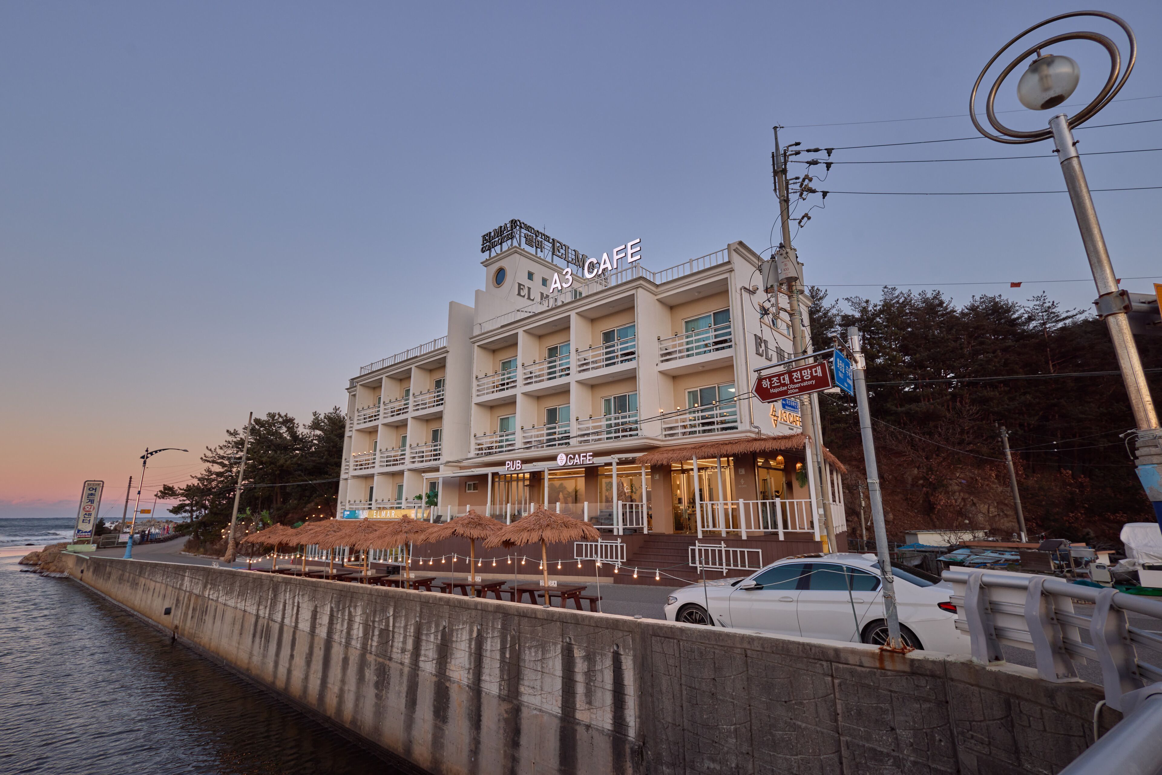 Um edifício de vários andares com café e pub no andar térreo, com mesas ao ar livre protegidas por guarda-sóis, vista para o mar e área de estacionamento.