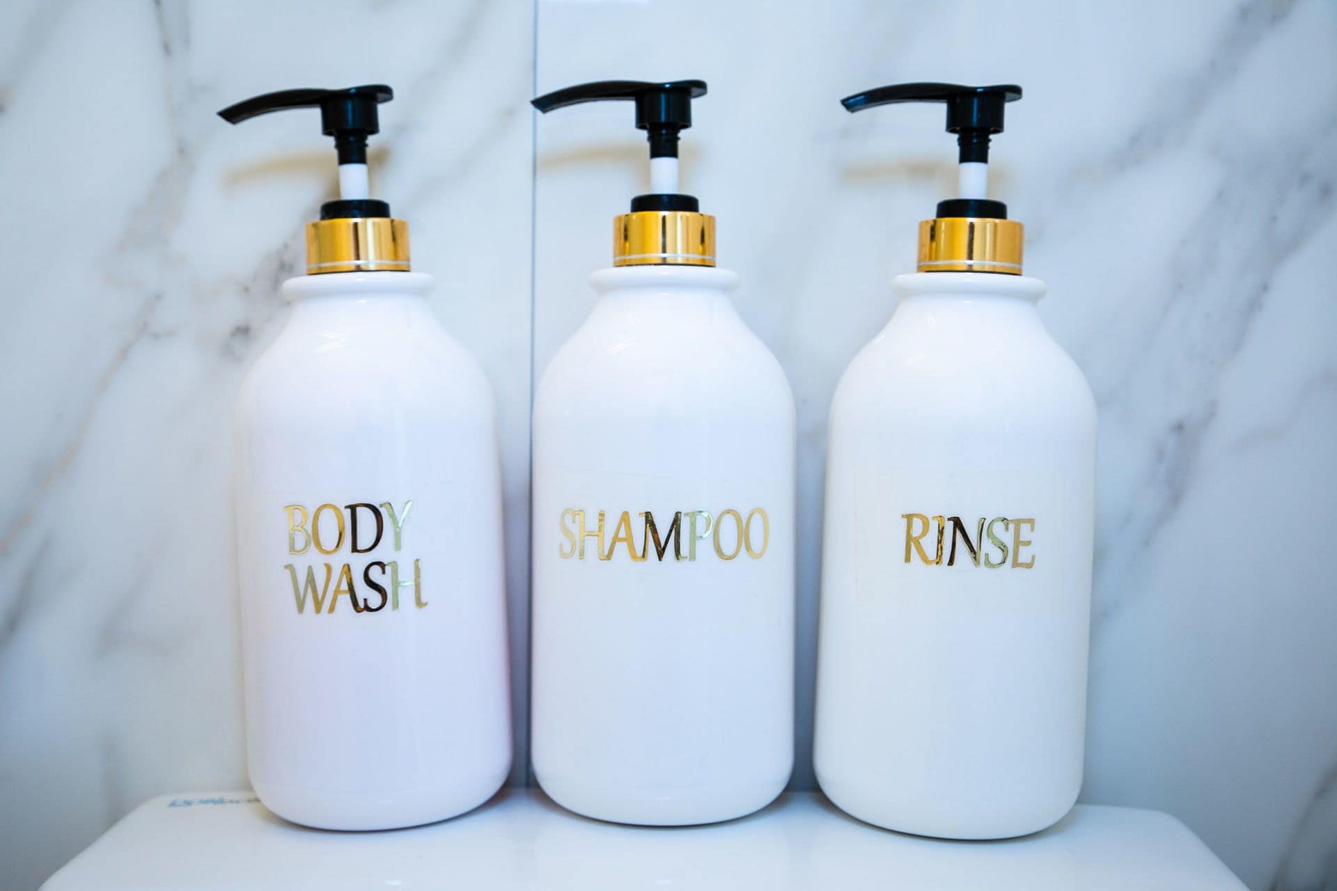 Three white bottles with gold and black pumps labeled "BODY WASH," "SHAMPOO," and "RINSE."