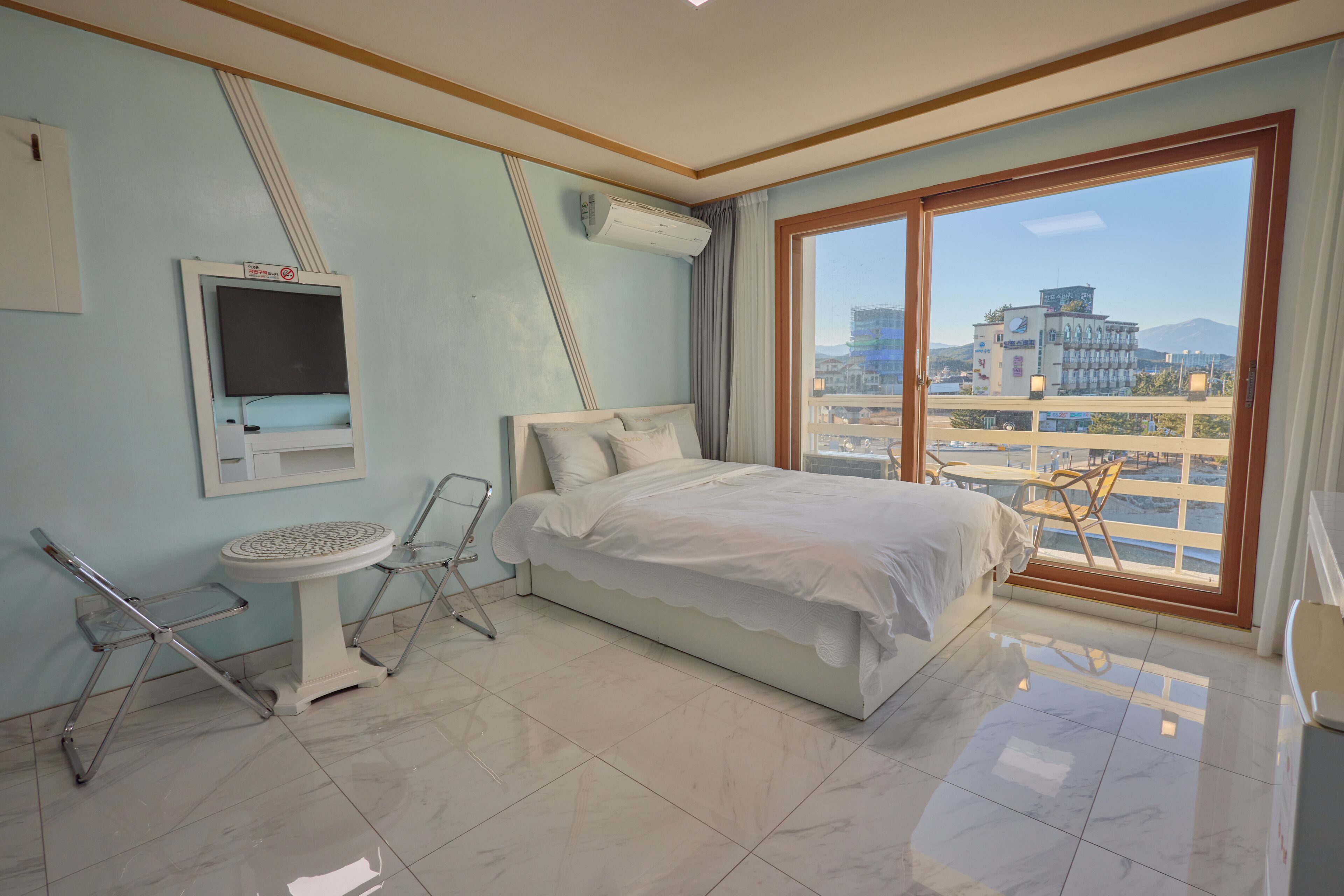 Ein Hotelzimmer mit einem Bett, einem kleinen Tisch, einem Stuhl, einer Klimaanlage und einem Fenster mit Blick auf Gebäude und einen Berg.