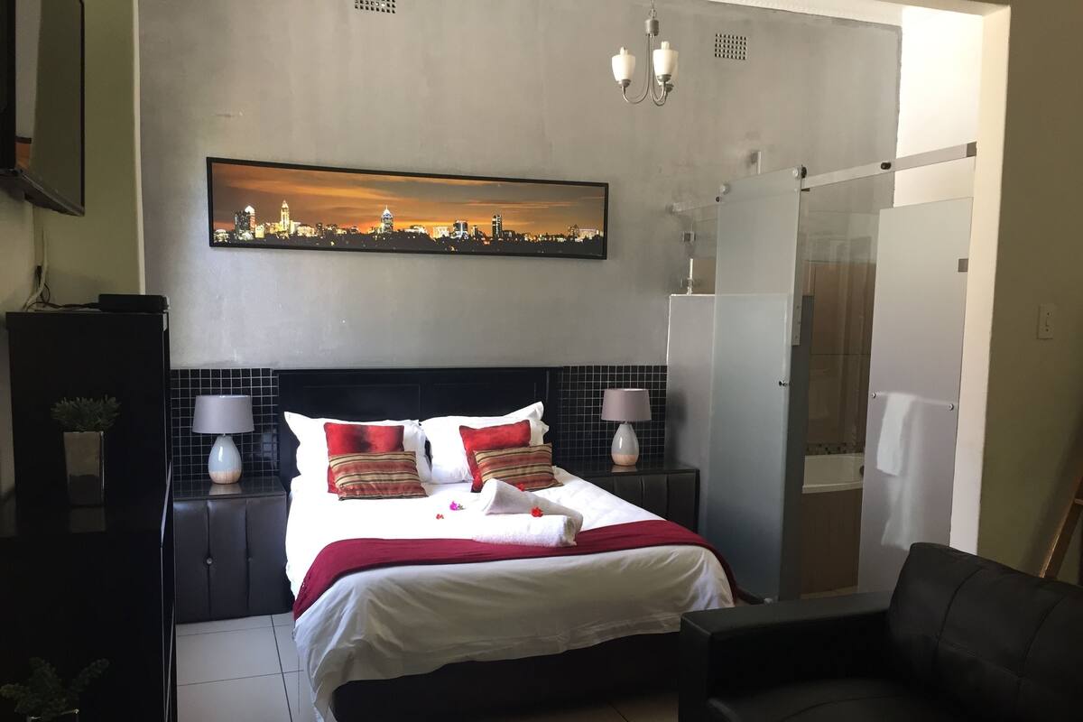 A hotel room with a bed, two bedside tables with lamps, a cityscape painting above the bed, a shower area, and a seating area with a chair.