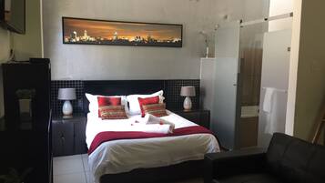 A hotel room with a bed, two bedside tables with lamps, a cityscape painting above the bed, a shower area, and a seating area with a chair.