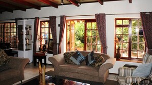 Ein gemütliches Wohnzimmer mit einer Couch, Sesseln und einem Couchtisch. Große Fenster mit Vorhängen, ein Bücherregal und eine dekorative Vase sind ebenfalls vorhanden.