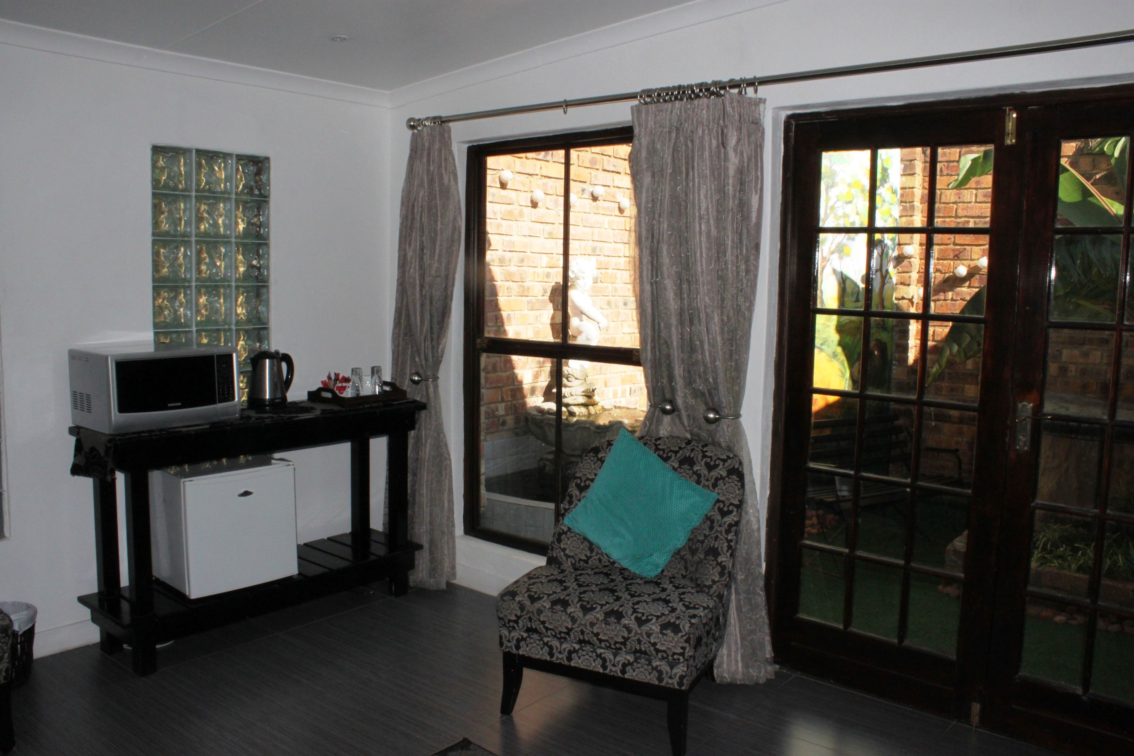 A compact room with a small white refrigerator, a microwave, and a coffee maker on a black table. A patterned chair with a teal pillow is positioned near a large window with curtains.