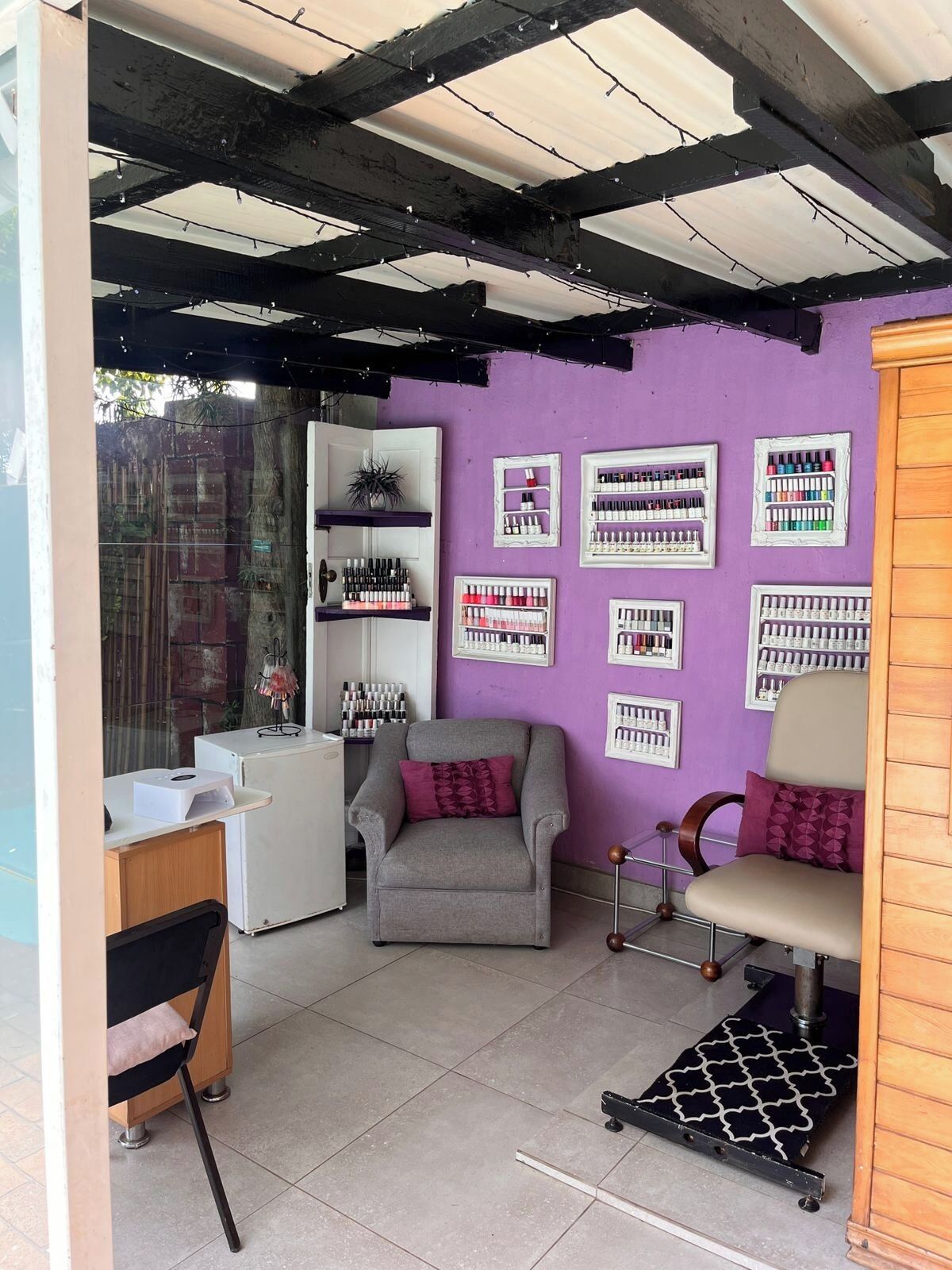 Interior de salón de belleza con pared morada, varios mostradores de esmalte de uñas, un sillón gris y un mueble de madera.