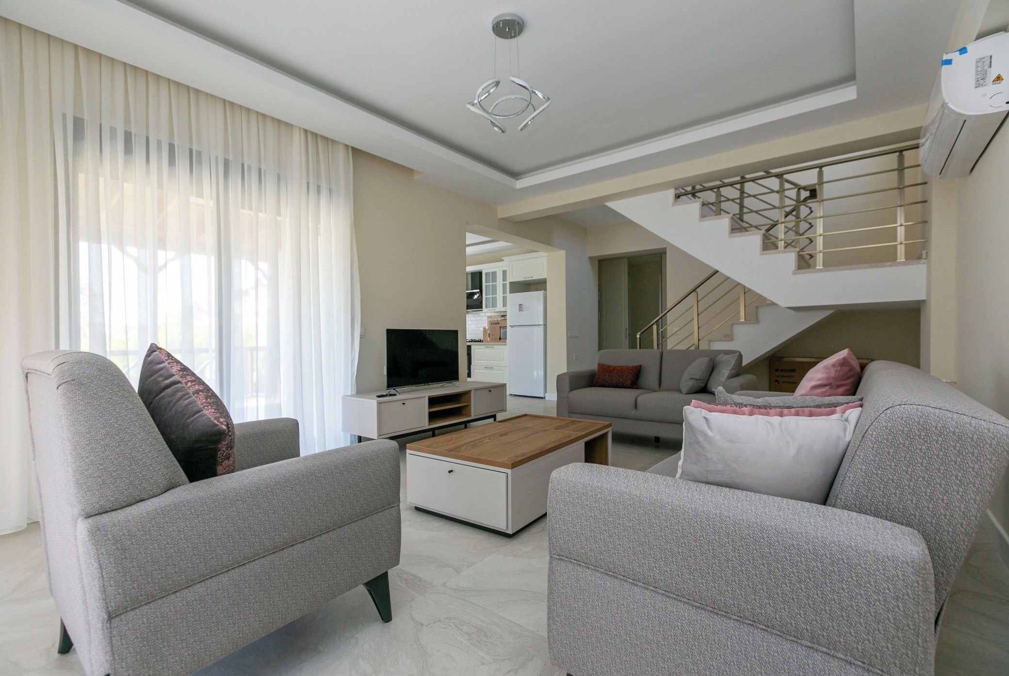 A modern living room with a sofa, coffee table, and TV. A staircase is visible in the background.