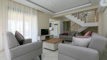 A modern living room with a sofa, coffee table, and TV. A staircase is visible in the background.