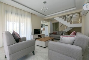 Un salon moderne avec un canapé, une table basse et un téléviseur. On aperçoit un escalier au fond.