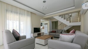 A modern living room with a sofa, coffee table, and TV. A staircase is visible in the background. - Paradise Town Villa Lisa (Serik)