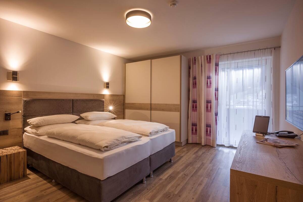 A bedroom with a bed, bedside tables, a desk with a computer, and a window with curtains.