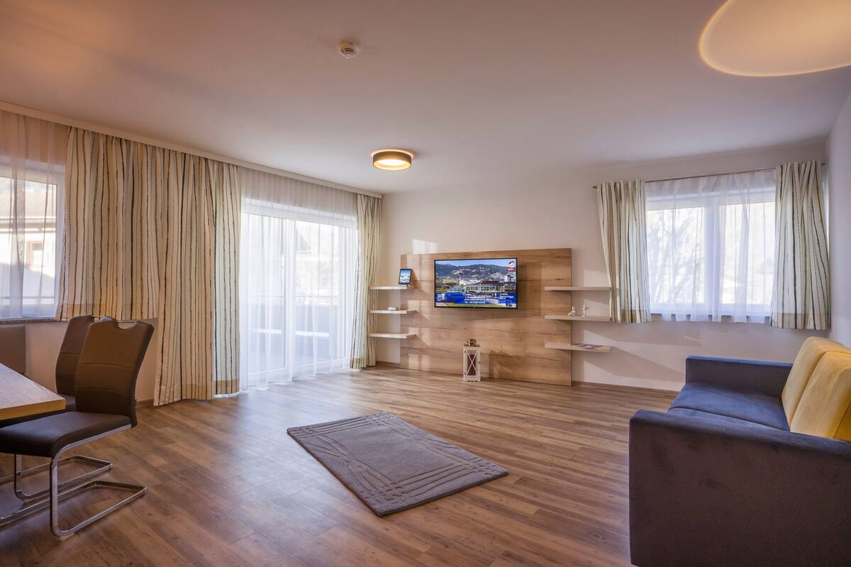 A modern living room with a wooden floor, a flat-screen TV mounted on the wall, a sofa, a chair, and a small table.