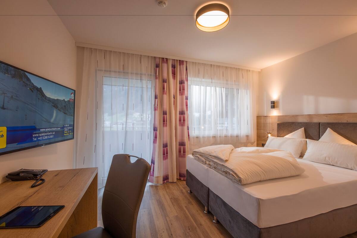 A hotel room with a bed, a desk with a phone and tablet, and a large TV on the wall.