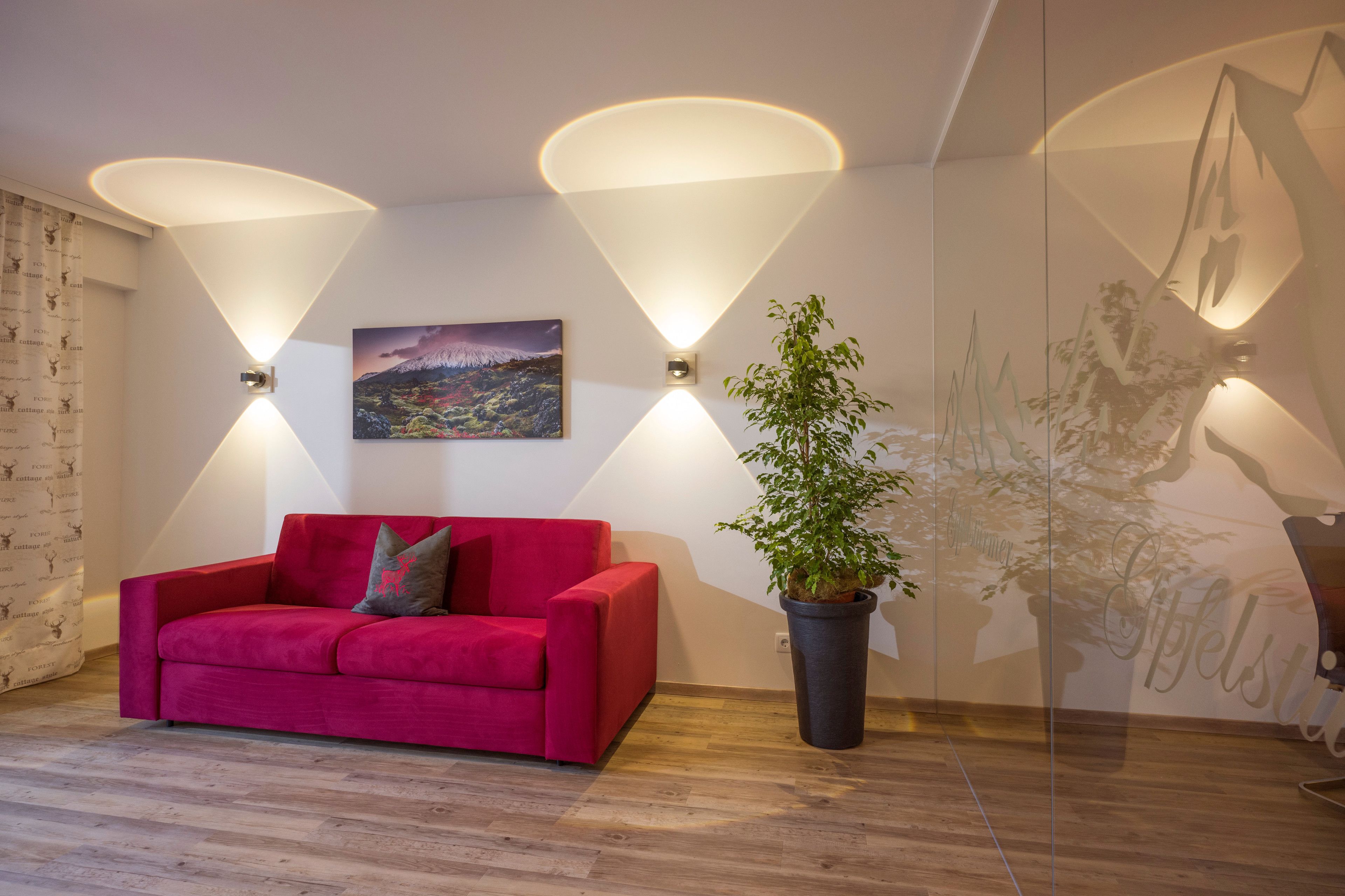 Een moderne kamer met een rode bank, twee wandlampen, een potplant en een stadsgezicht schilderij.