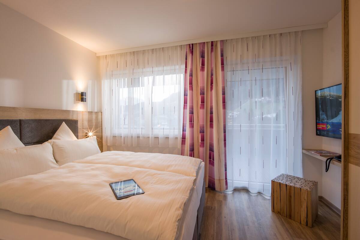 A hotel room with a large bed, a bedside table, a TV, and a window with sheer curtains.