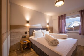 A modern hotel room with a large bed, wooden headboard, two wall-mounted lights, a small wooden side table, and a window with curtains.