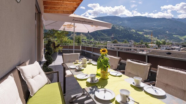 Una terrazza con tavolo apparecchiato per mangiare, una pianta in vaso e vista sulle montagne.