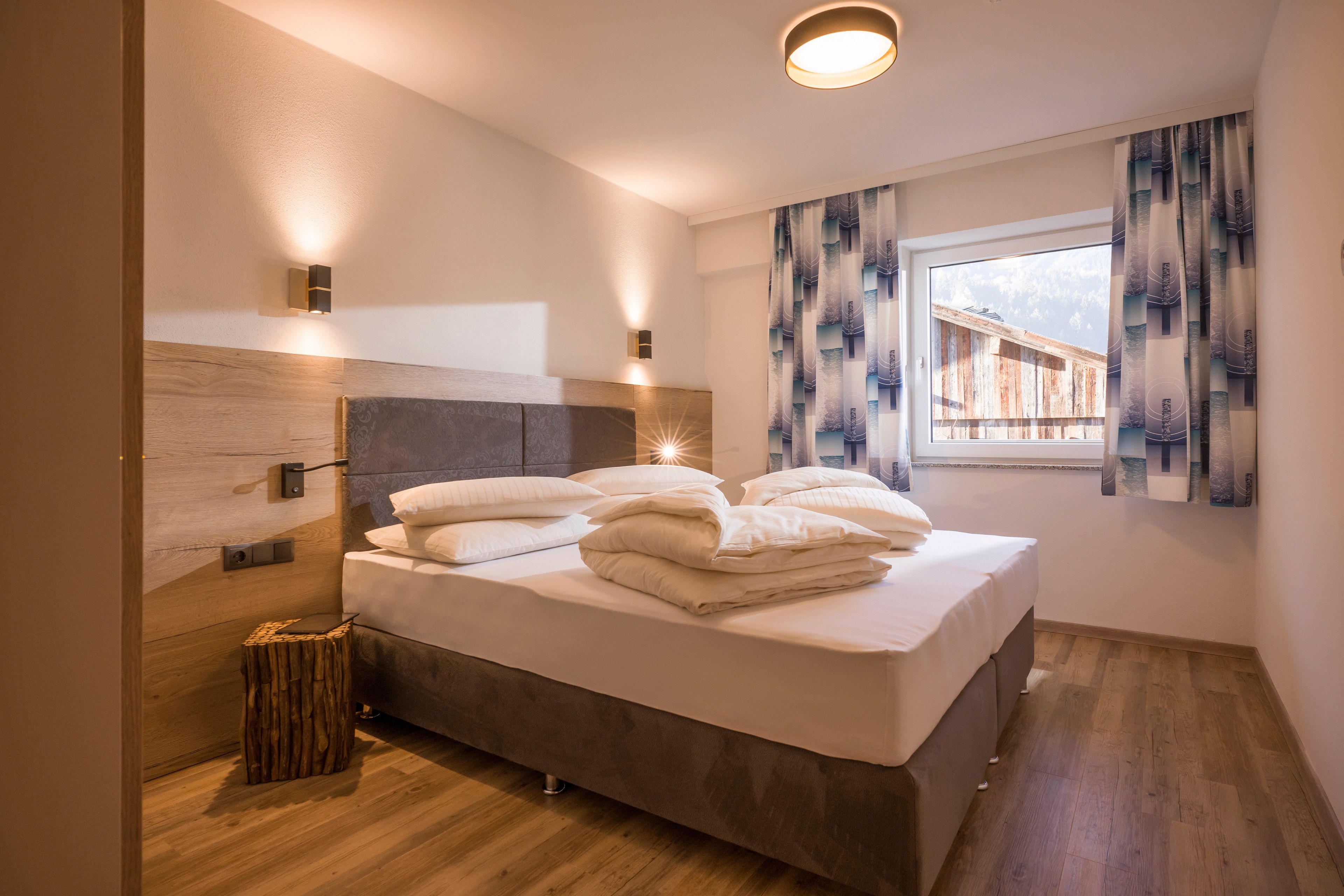 A modern bedroom with a wooden headboard, a large bed with neatly folded white linens, a small wooden stool, and a window with patterned curtains.