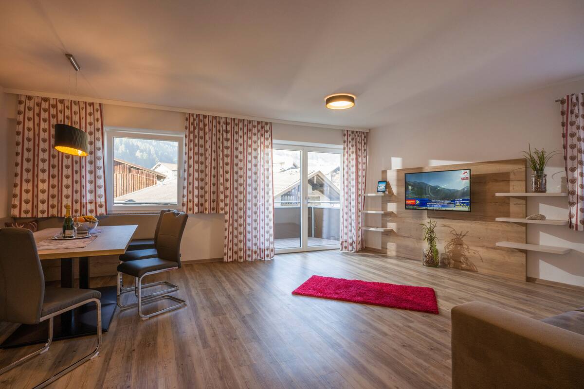 A modern living room with a dining area, a TV mounted on the wall, and a large window with curtains.