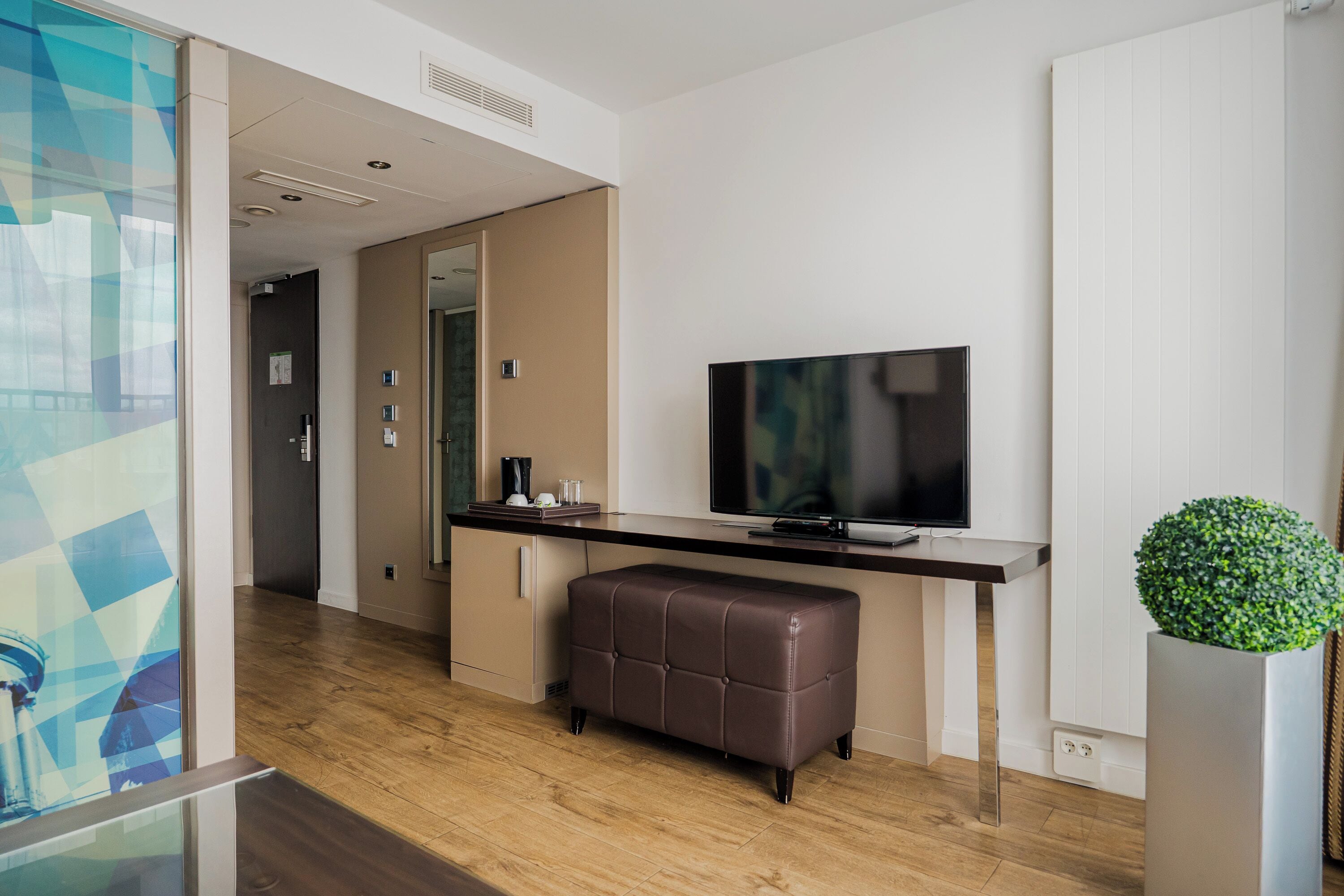 Une chambre d’hôtel moderne avec un plancher en bois, un téléviseur à écran plat, un banc en cuir marron et une plante en pot.