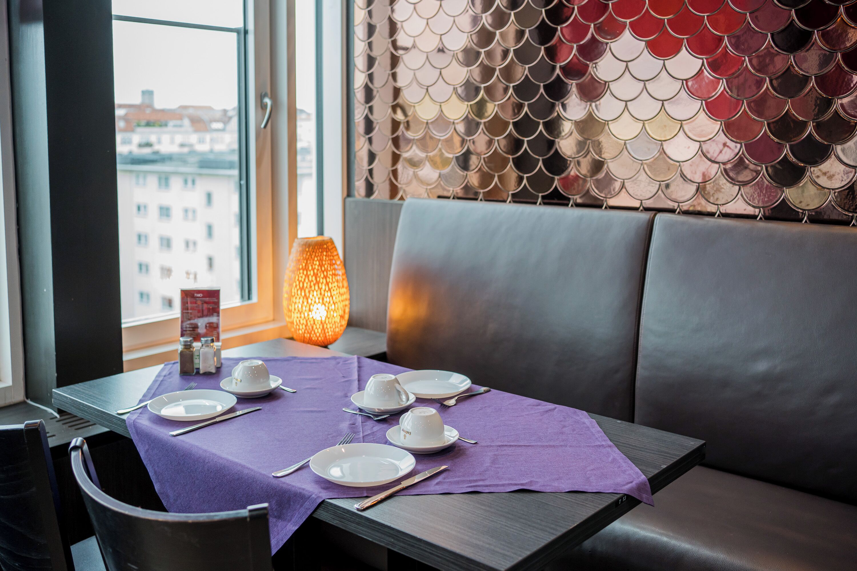 A dining area with a table set for four, a purple tablecloth, and a view of buildings through a window.