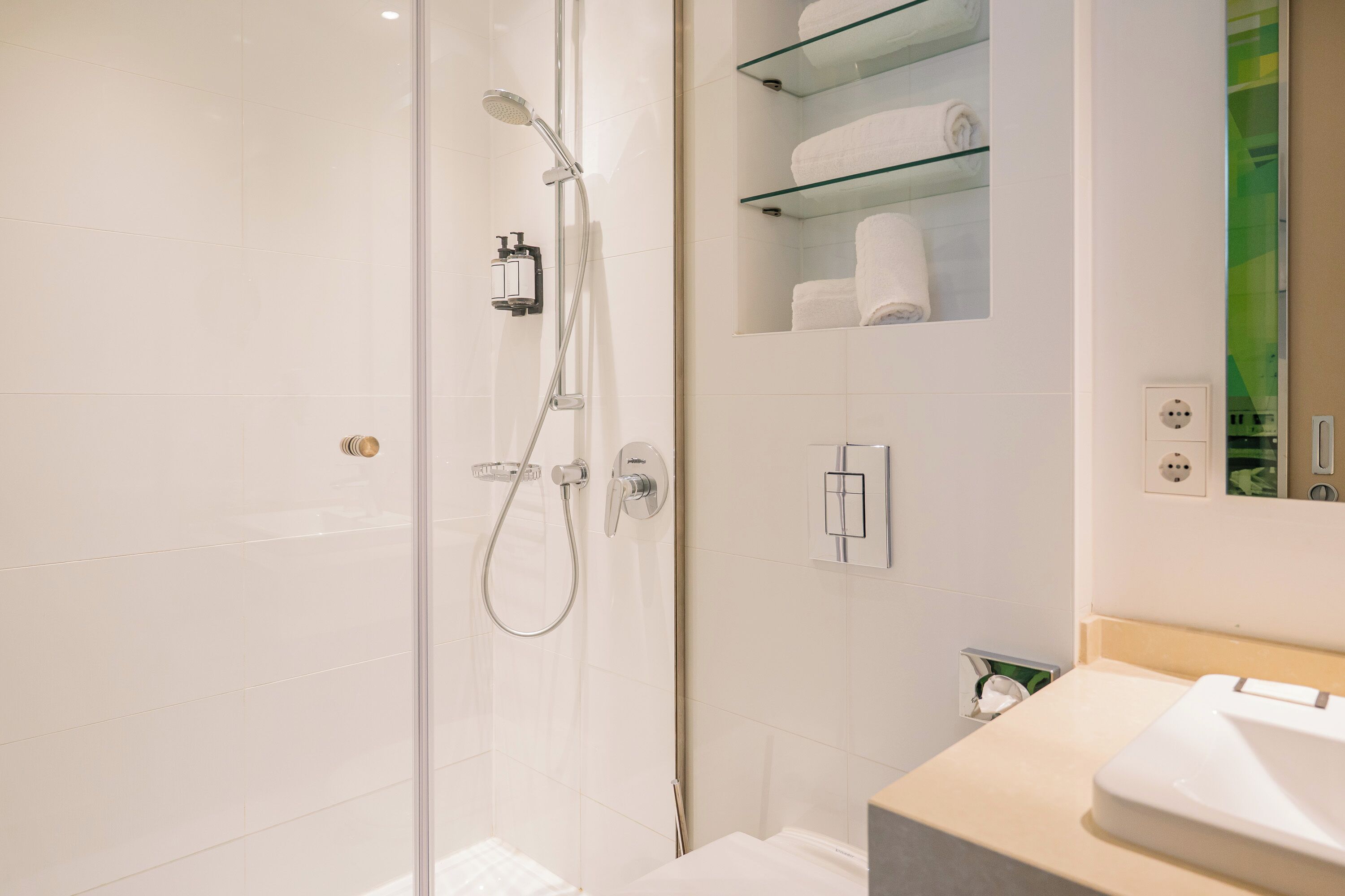 Ein modernes Badezimmer mit einer Duschkabine aus Glas, einem weißen Waschbecken und Glastregalen mit Handtüchern.