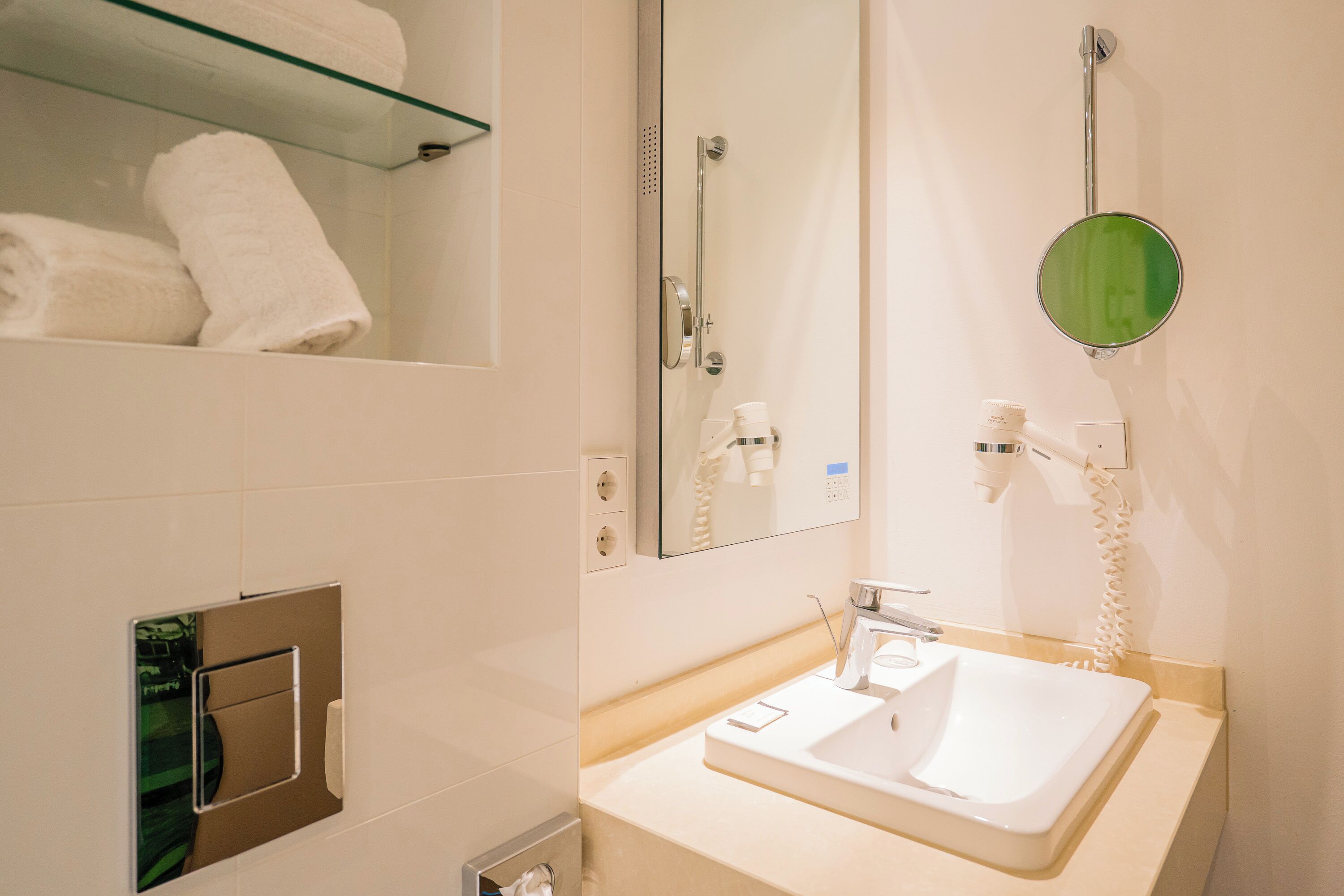 A modern bathroom with a glass shelf holding towels, a large mirror, a sink with a faucet, and a handheld showerhead.