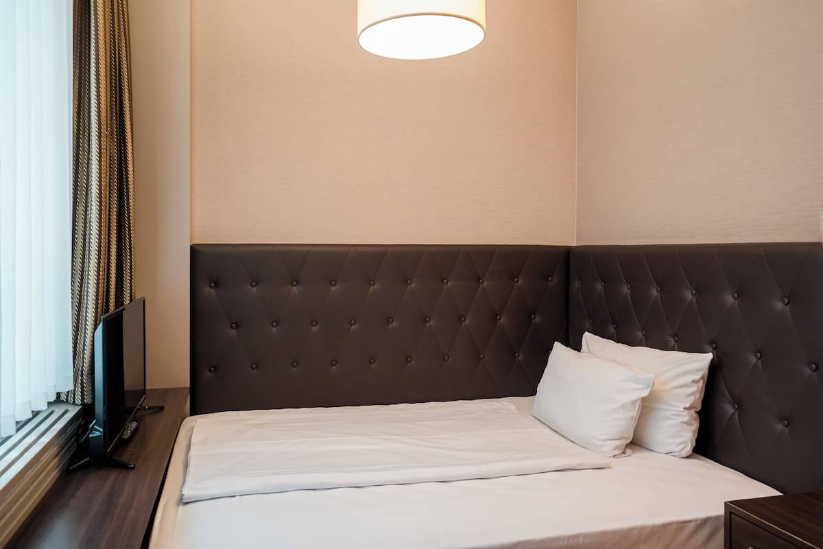 Een netjes opgemaakt bed met witte lakens, een donker gestoffeerd hoofdbord en een televisie op een houten nachtkastje.