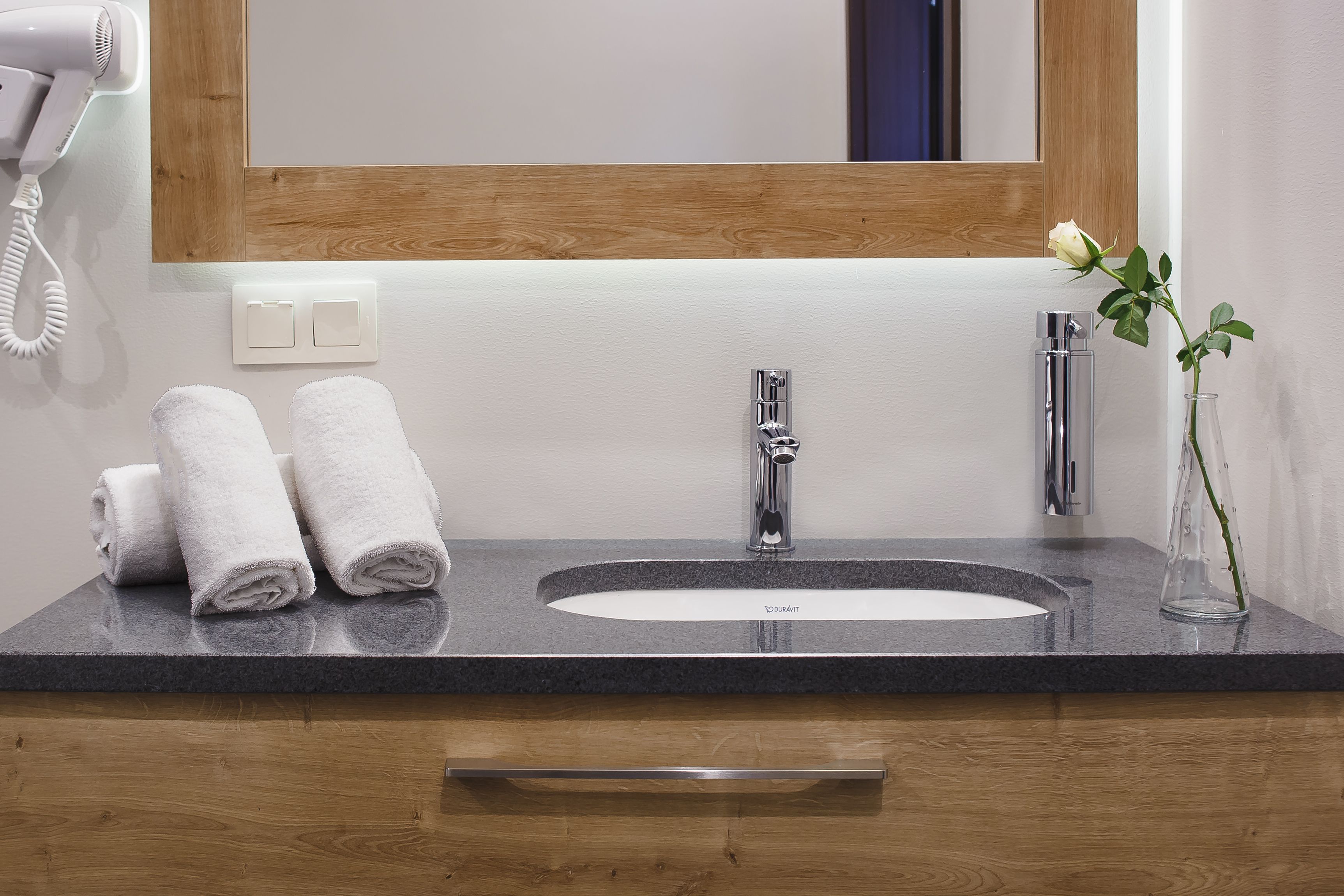 A bathroom with a granite countertop, a white sink, a mirror, a hairdryer, and a vase with a single rose.