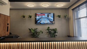 Un área de recepción con diseño moderno, que incluye un mostrador de madera, un televisor de pantalla plana montado en la pared y varias plantas colgantes.