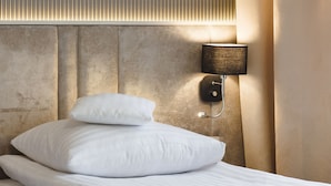 Una cama bien hecha con sábanas y almohadas blancas, una lámpara de pared y un panel de pared texturizado.