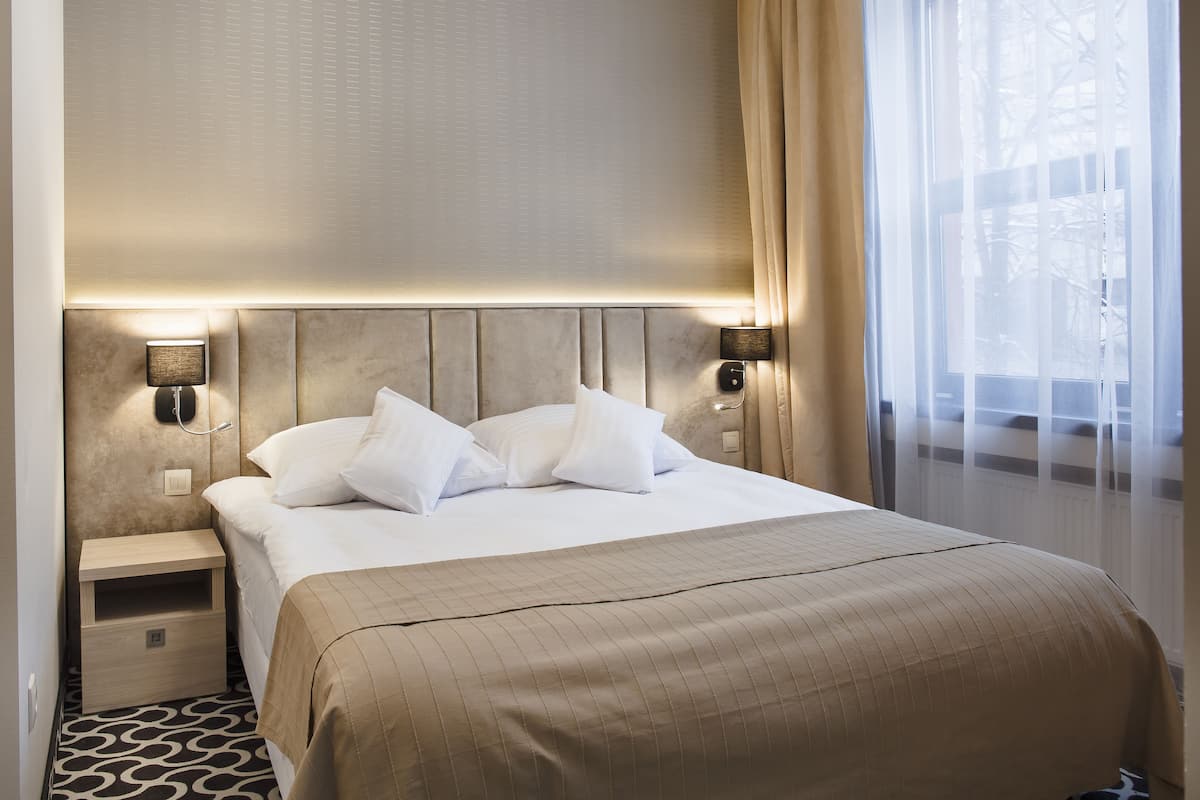 Habitación de hotel con una cama grande, dos mesitas de noche, cabecera con iluminación integrada y una ventana con cortinas translúcidas.