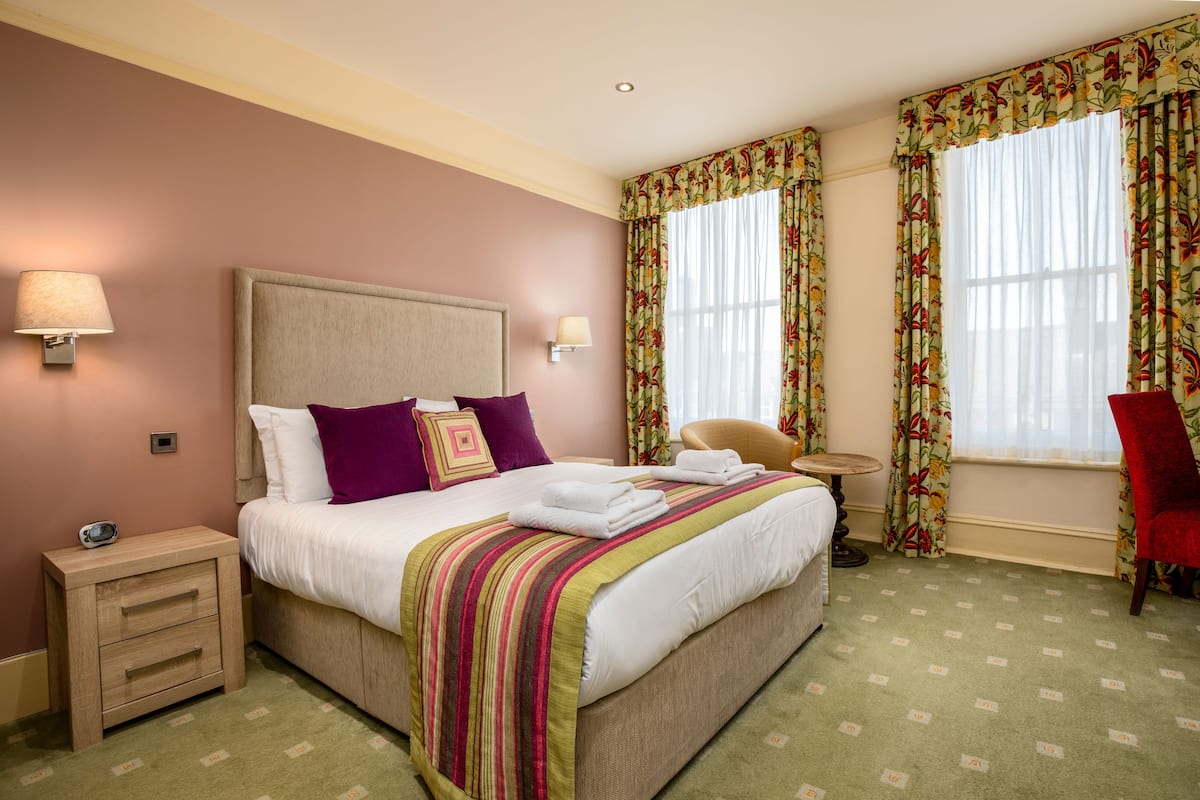 Ein Hotelzimmer mit einem großen Bett, zwei Nachttischlampen, einem Nachttisch, einem Sessel, einem kleinen Tisch und blumigem Vorhang.