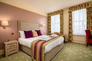 Quarto de hotel com uma cama grande, duas luminárias de cabeceira, uma mesinha de cabeceira, uma cadeira, uma pequena mesa e cortinas florais.