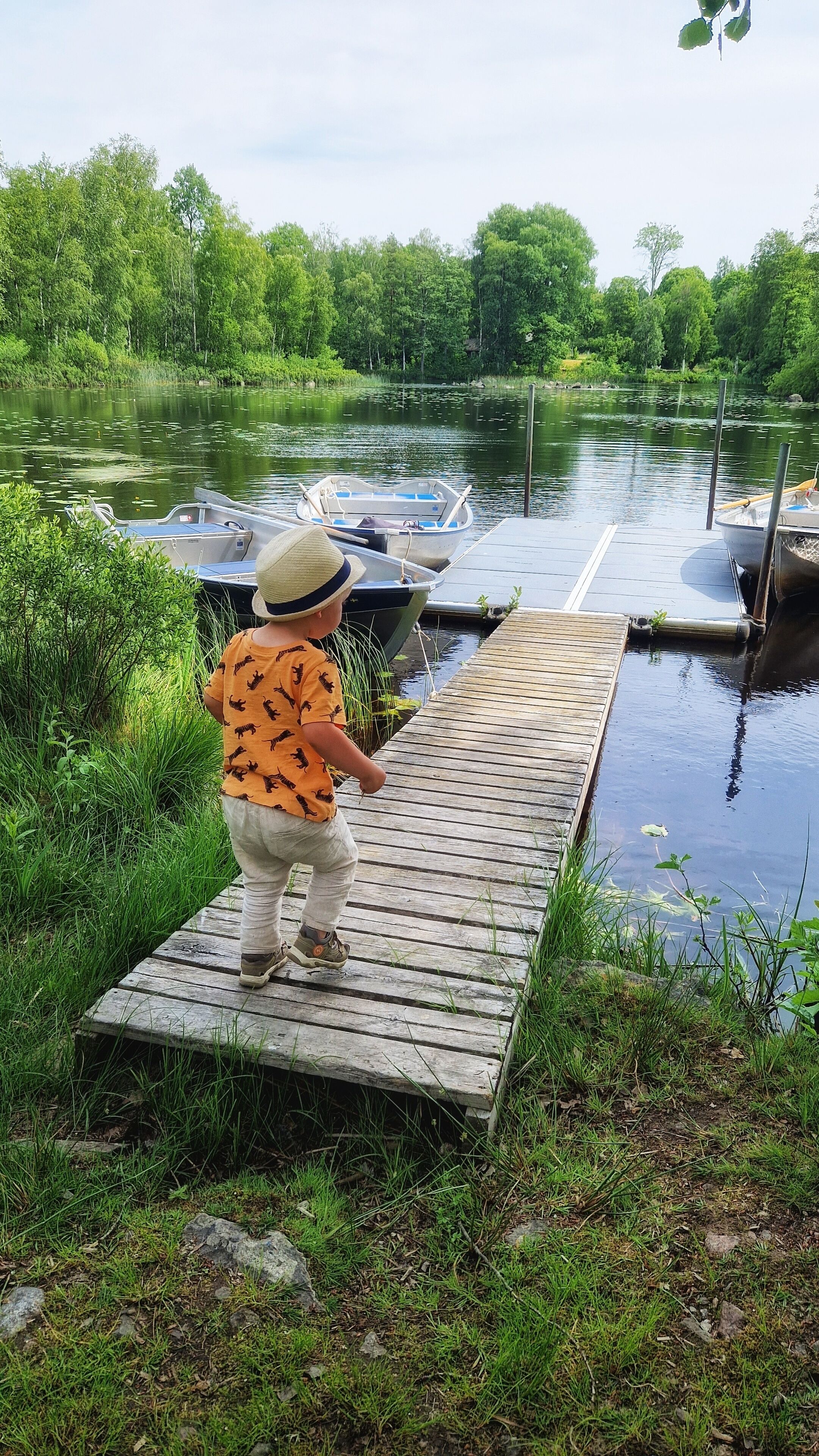 Un enfant se tient debout sur une jetée en bois, au bord d’un lac paisible, avec des bateaux amarrés à proximité.