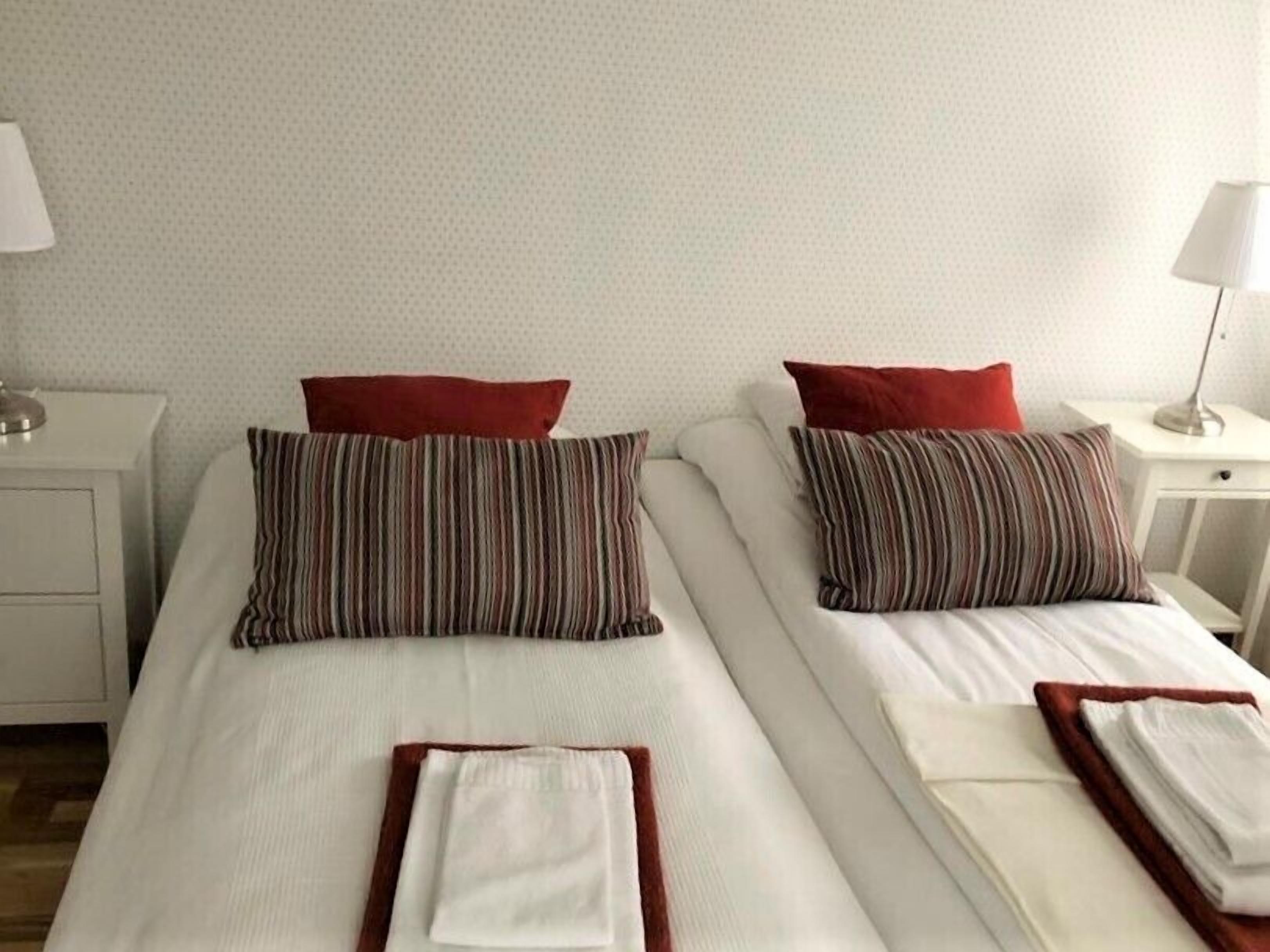 Deux lits simples avec des oreillers à rayures et des coussins rouges, du linge de lit blanc et une petite table de chevet avec une lampe.