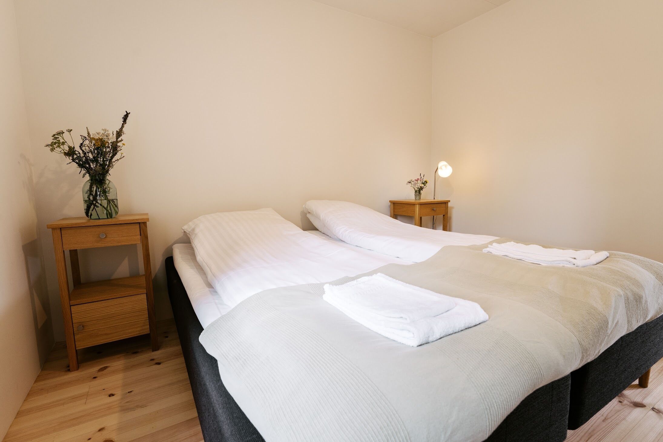 A neatly made bed with white linens and a folded towel, two bedside tables with lamps, and a vase with dried flowers on one table.