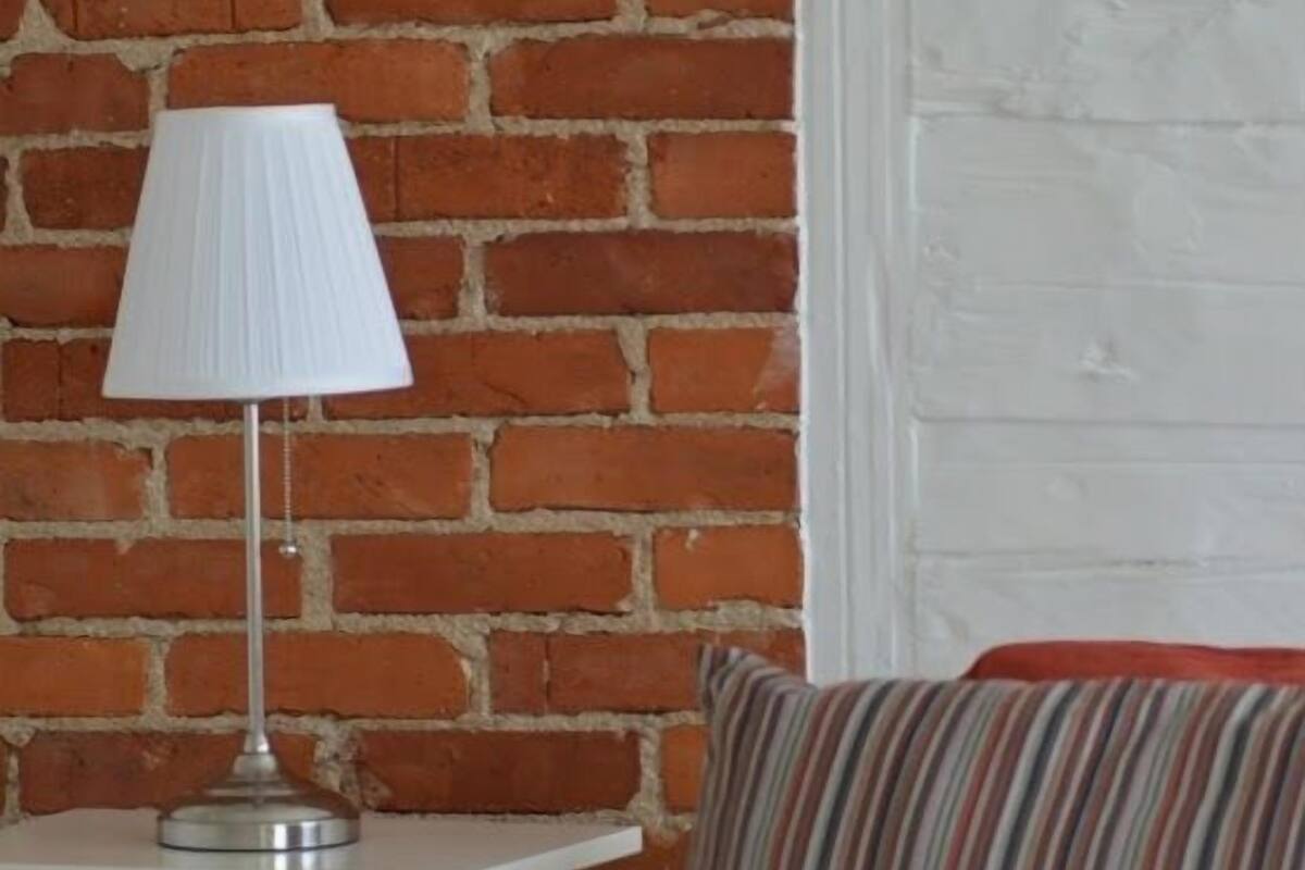 A room with a brick wall, a white bedside table with a lamp, a striped pillow, and a bed with a red blanket.