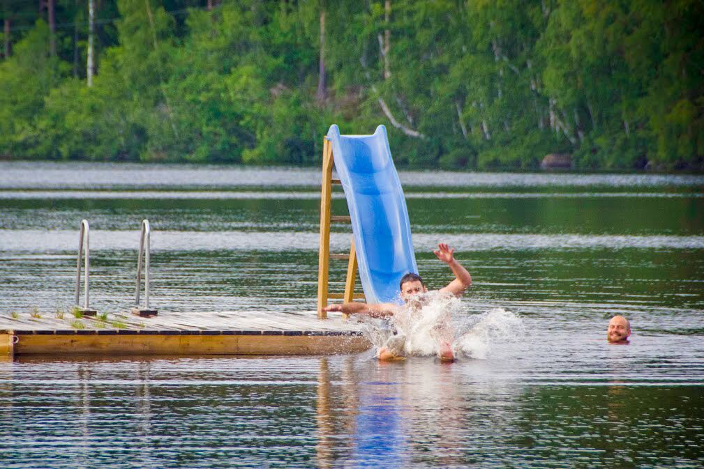 Un hombre se desliza por un tobogán azul hacia el agua, con un muelle y follaje al fondo.