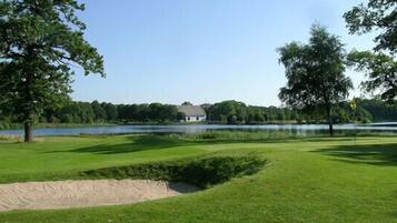 Campo de golf con un búnker de arena, un estanque y una casa al fondo.
