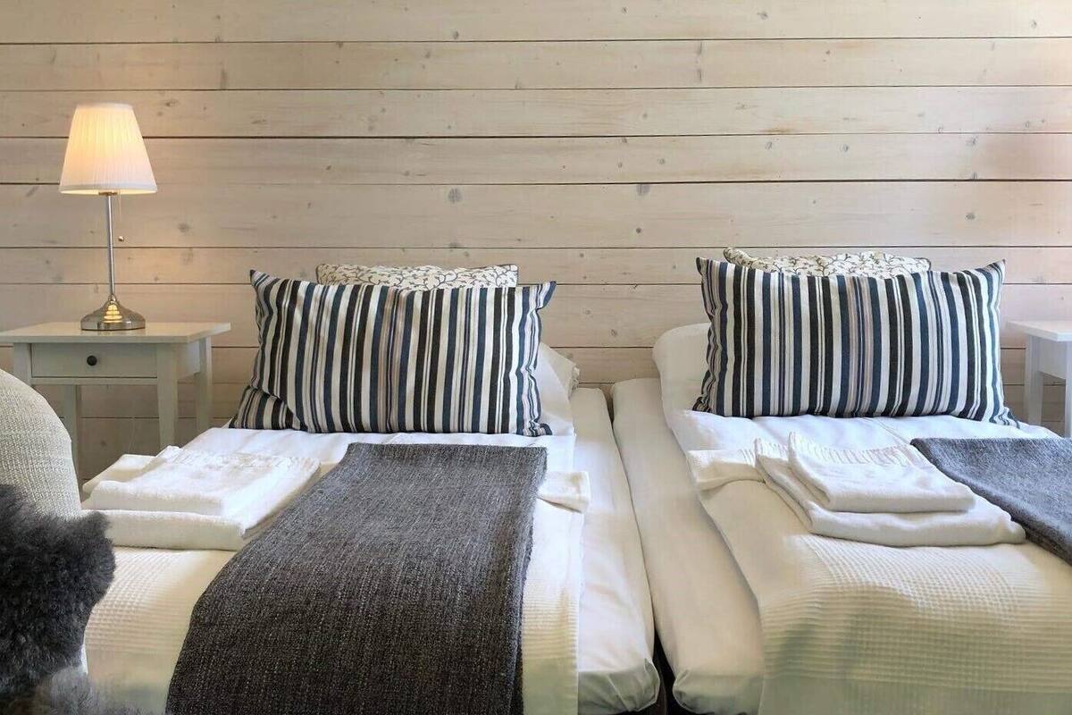 A double room with two beds, each with striped pillows and a blanket, a bedside table with a lamp, and a wooden wall.