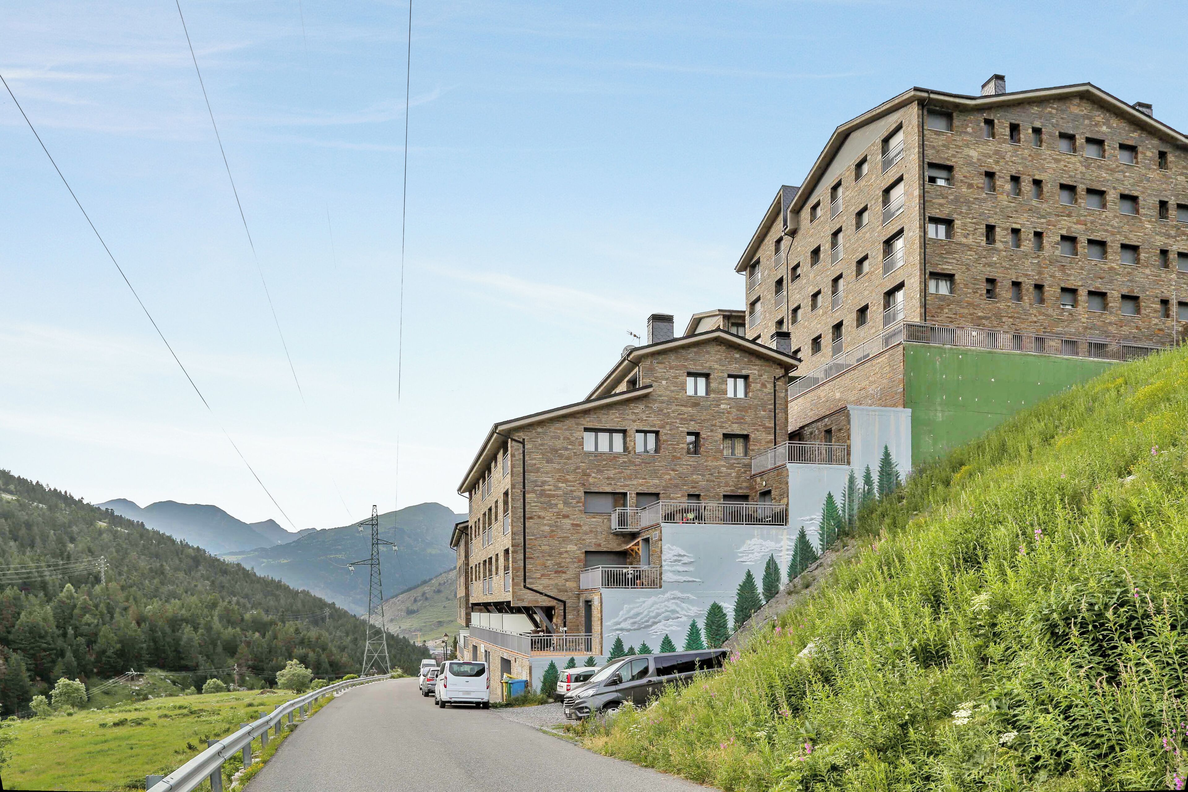 Un edificio de varios pisos con fachada de piedra, una ladera cubierta de vegetación y una calle con coches estacionados.