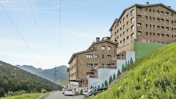 Un edificio de varios pisos con fachada de piedra, una ladera cubierta de vegetación y una calle con coches estacionados.