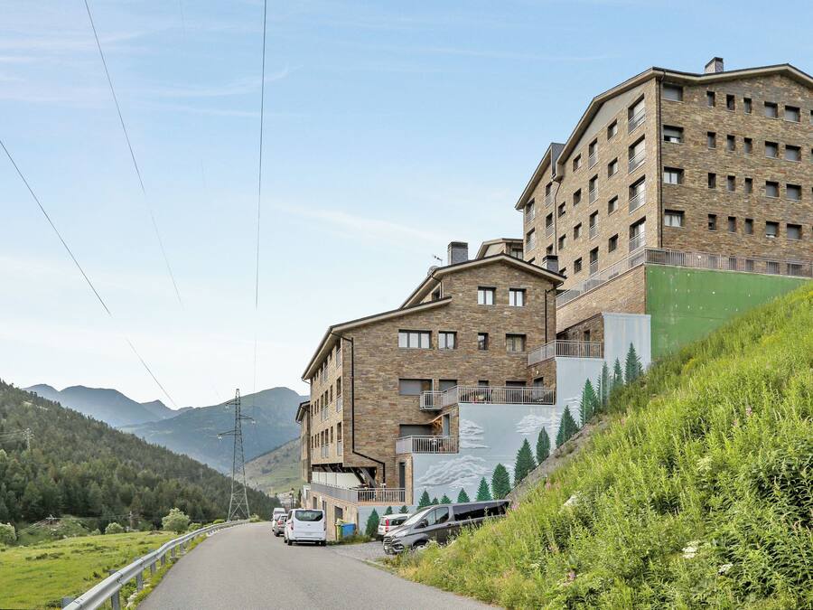 Un edificio de varios pisos con fachada de piedra, una ladera cubierta de vegetación y una calle con coches estacionados.