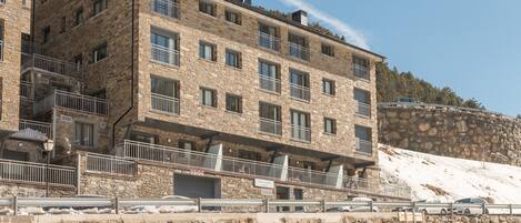Un edificio de varios pisos de piedra con balcones y fachada de piedra, y un área de estacionamiento al frente.