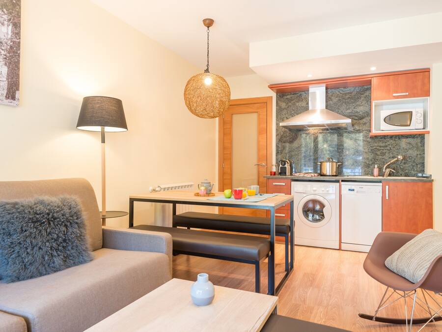 En kompakt opholdsstue med sofa, spisebord, tekøkken med mikrobølgeovn, vaskemaskine og en hængende lampe.